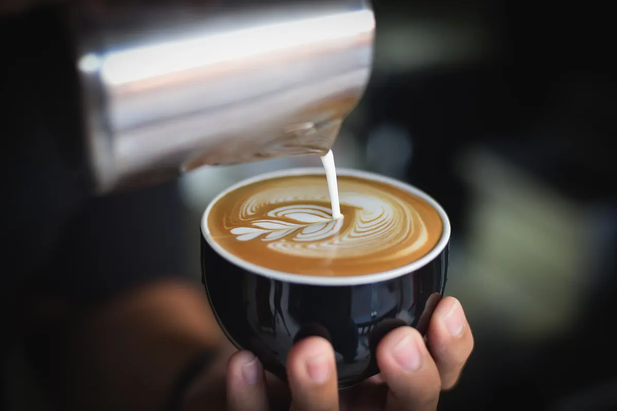 Jarra de acero vertiendo leche para dibujar arte latte en una taza oscura